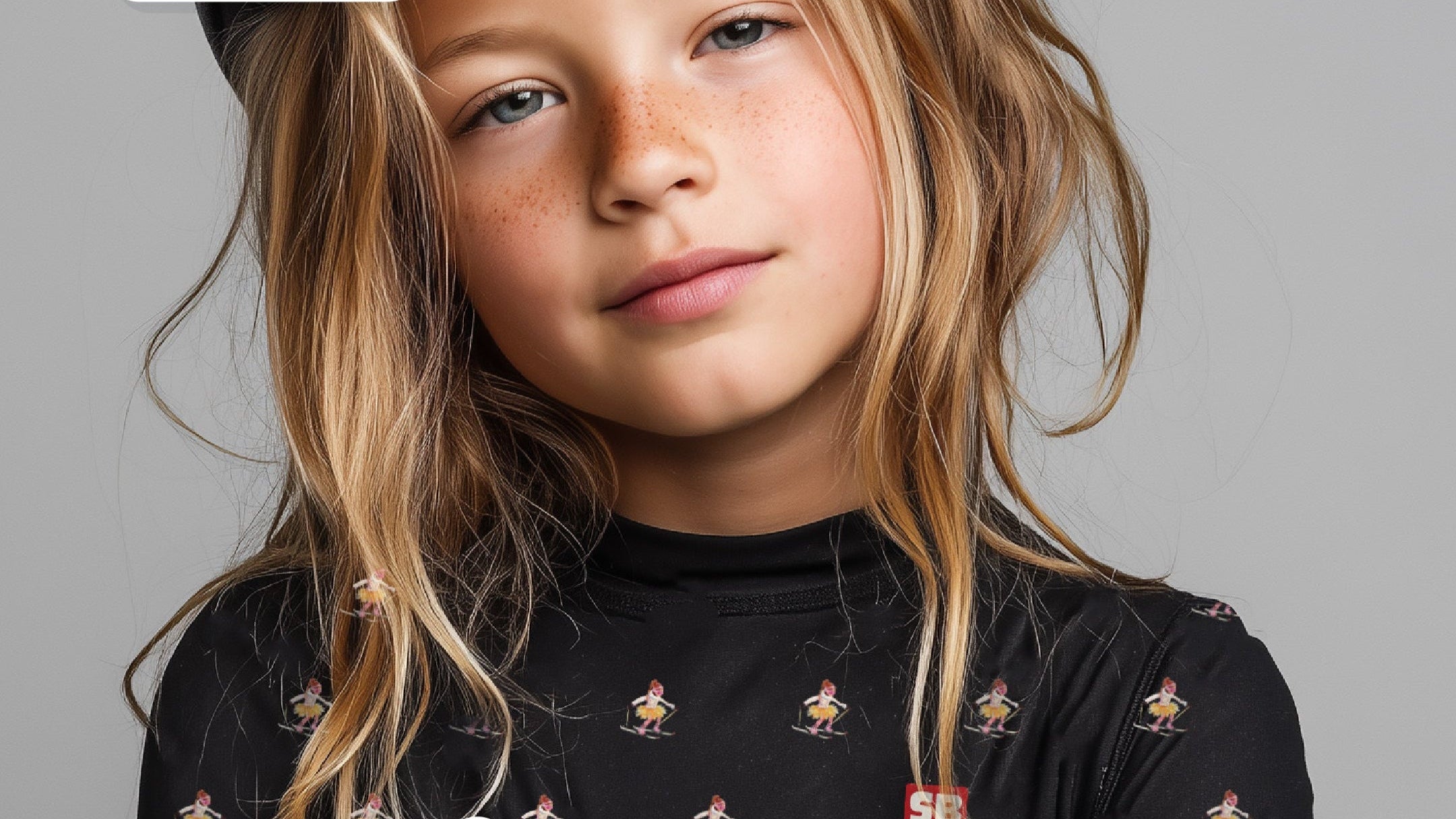 Child wearing a black long-sleeve shirt with a graphic design, helmet, and goggles on a gray background. SB Baselayers logo is visible.